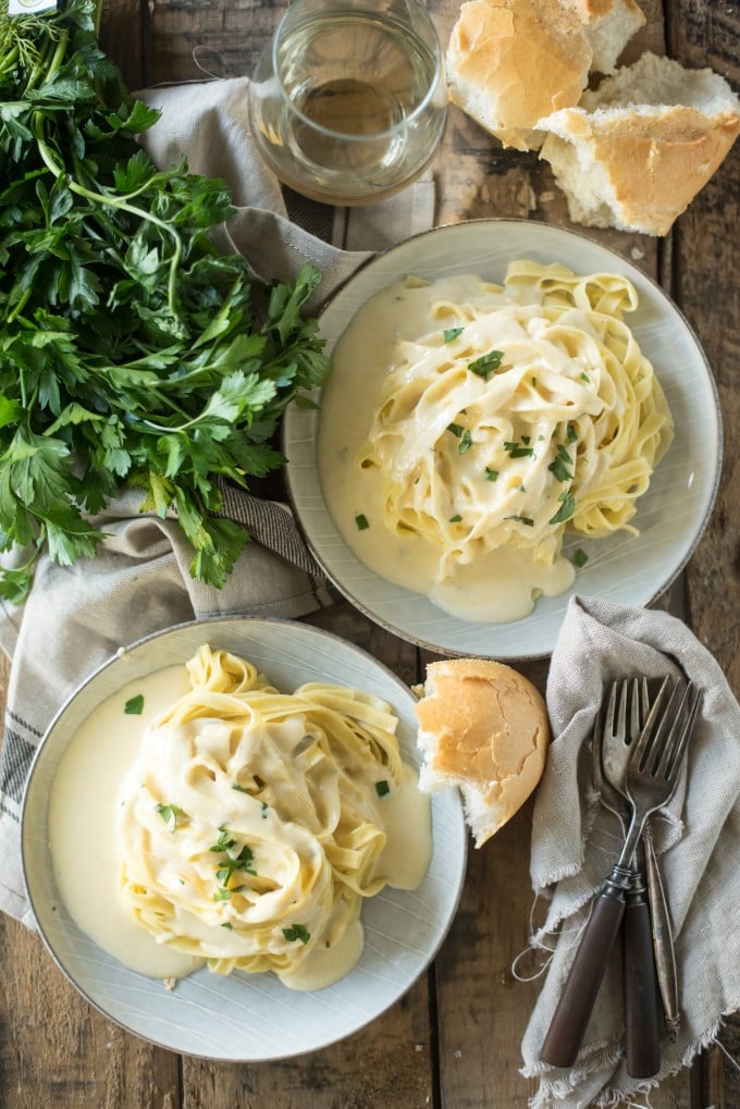 Overhead view of two plates of fettuccine Alfredo on gray plates on wood table with forks, bread and parsley