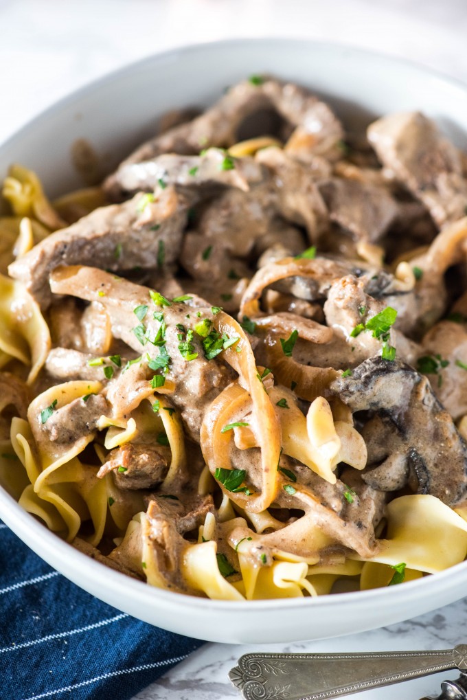 This easy homemade beef stroganoff is the best authentic recipe out there! Ready in under 30 minutes, you can use a variety of beef cuts depending on your budget. Beef Stroganoff over egg noodles in white bowl, close up shot