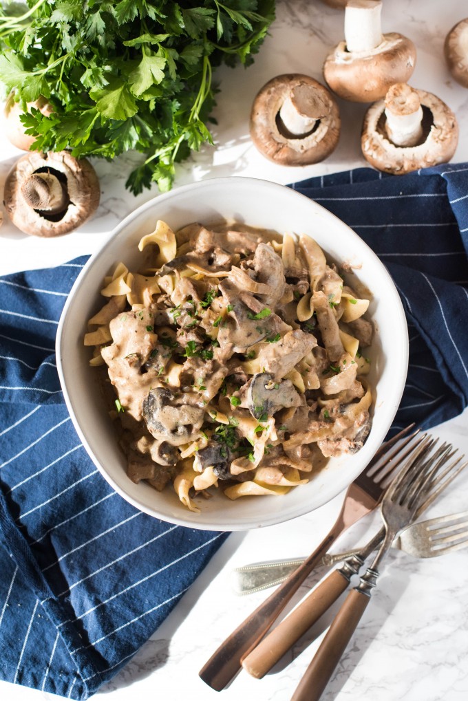 Best slow cooker beef stroganoff recipe with parsley, mushrooms and onions on a blue napkin in a white bowl with utensils