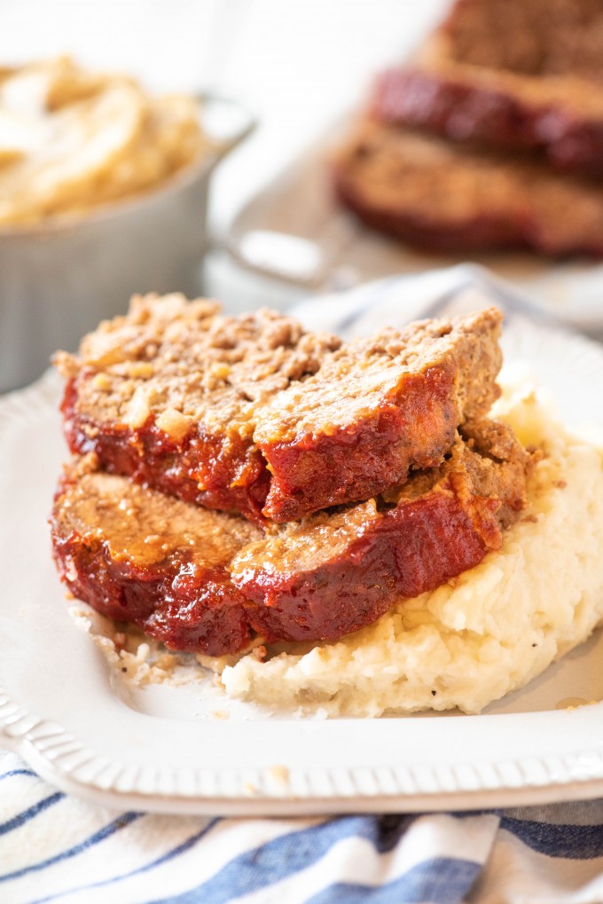 This easy Instant Pot Meatloaf and Mashed Potatoes recipe is the best! It's an easy way to make an entire comfort food meal in under an hour! After you've tried it, this will be the only meatloaf recipe you'll ever use! Pressure cooking has never been so easy! #gogogogourmet #instantpotmeatloaf #meatloafandmashedpotatoes #easyinstantpotrecipes #comfortfoodrecipes Two slices of instant pot meatloaf on top of mashed potatoes
